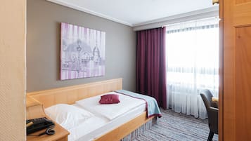 Ein Hotelzimmer mit einem hölzernen Bett, einem Schreibtisch mit Telefon, einem Stuhl, einem Fenster mit transparenten Vorhängen und einem an der Wand hängenden Kunstwerk.