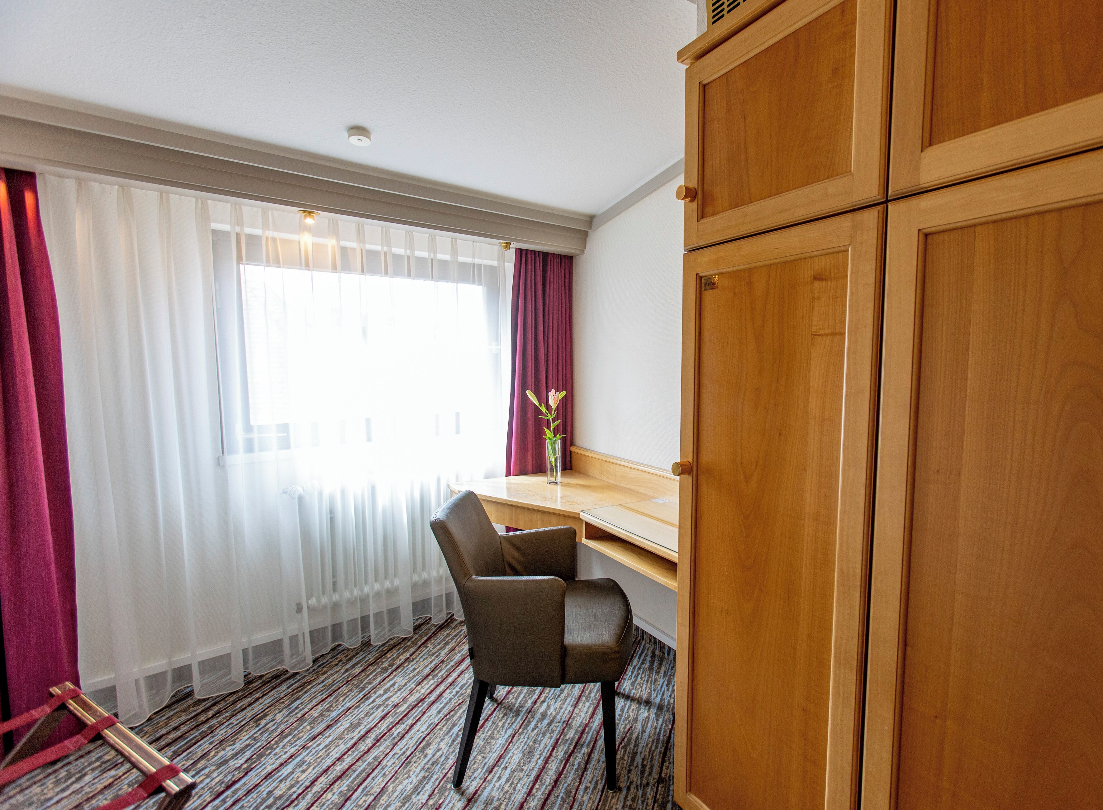 Habitación de hotel con un escritorio de madera, una silla, un ventanal con cortinas traslúcidas y un radiador.