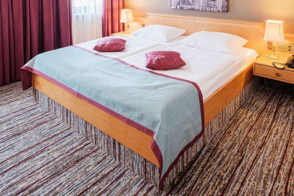 Ein Hotelzimmer mit einem großen Bett, zwei Nachttischlampen, einer Kopfteil und einem gemusterten Teppich, sowie einem Stadtbild an der Wand.