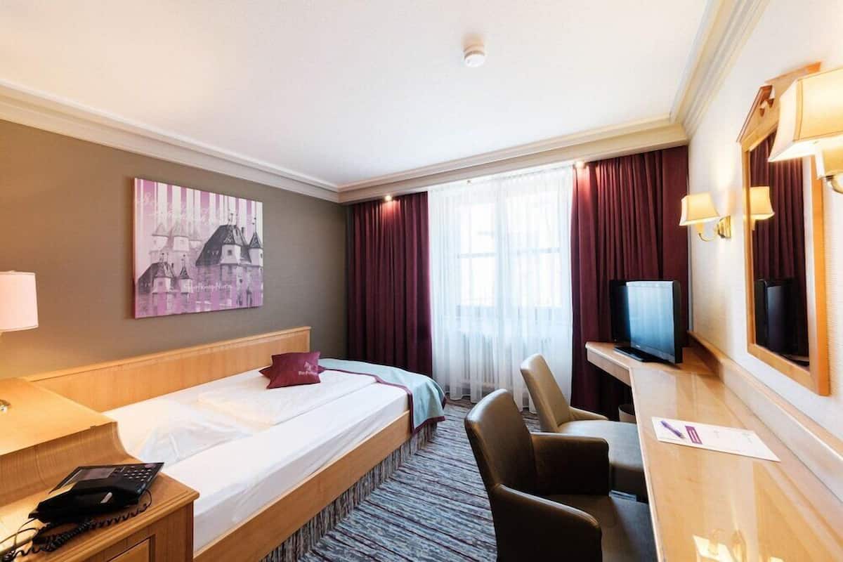 Ein Hotelzimmer mit Bett, Schreibtisch mit Computer, Stuhl, Fernseher und einem großen Fenster mit Vorhängen.
