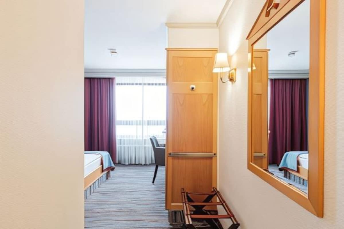 A hotel room with a bed, a desk, a chair, a wooden wardrobe, and a large mirror.
