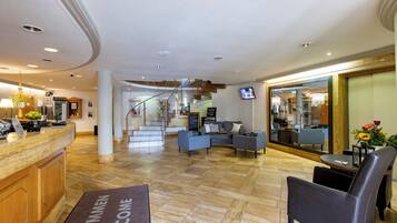 A hotel lobby with a reception desk, seating area, staircase, and a TV screen.