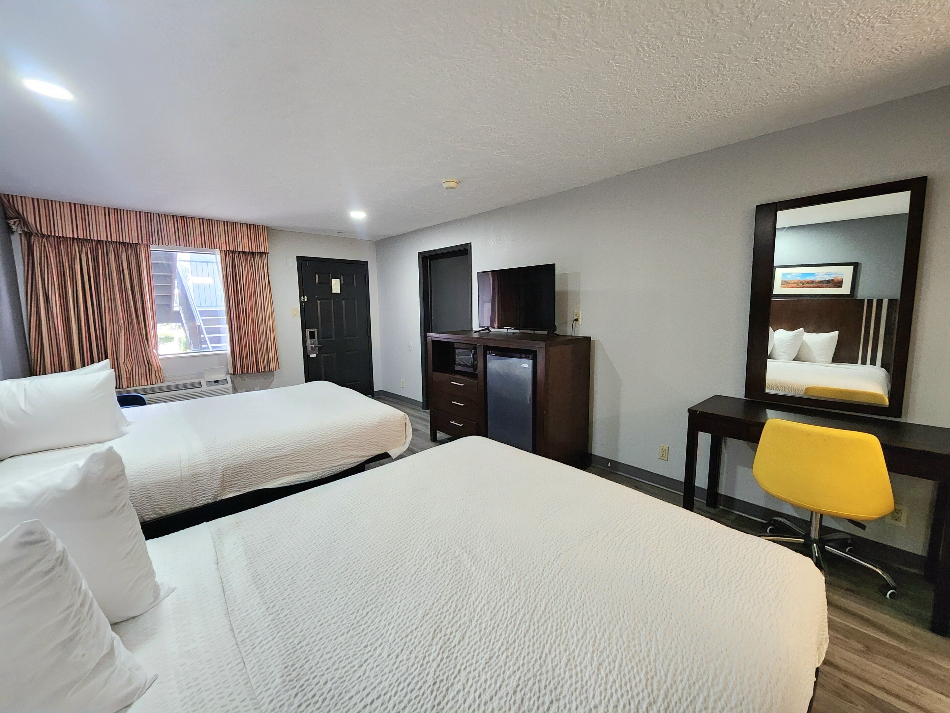 A hotel room with two beds, a desk with a television, a mirror, and a yellow chair.