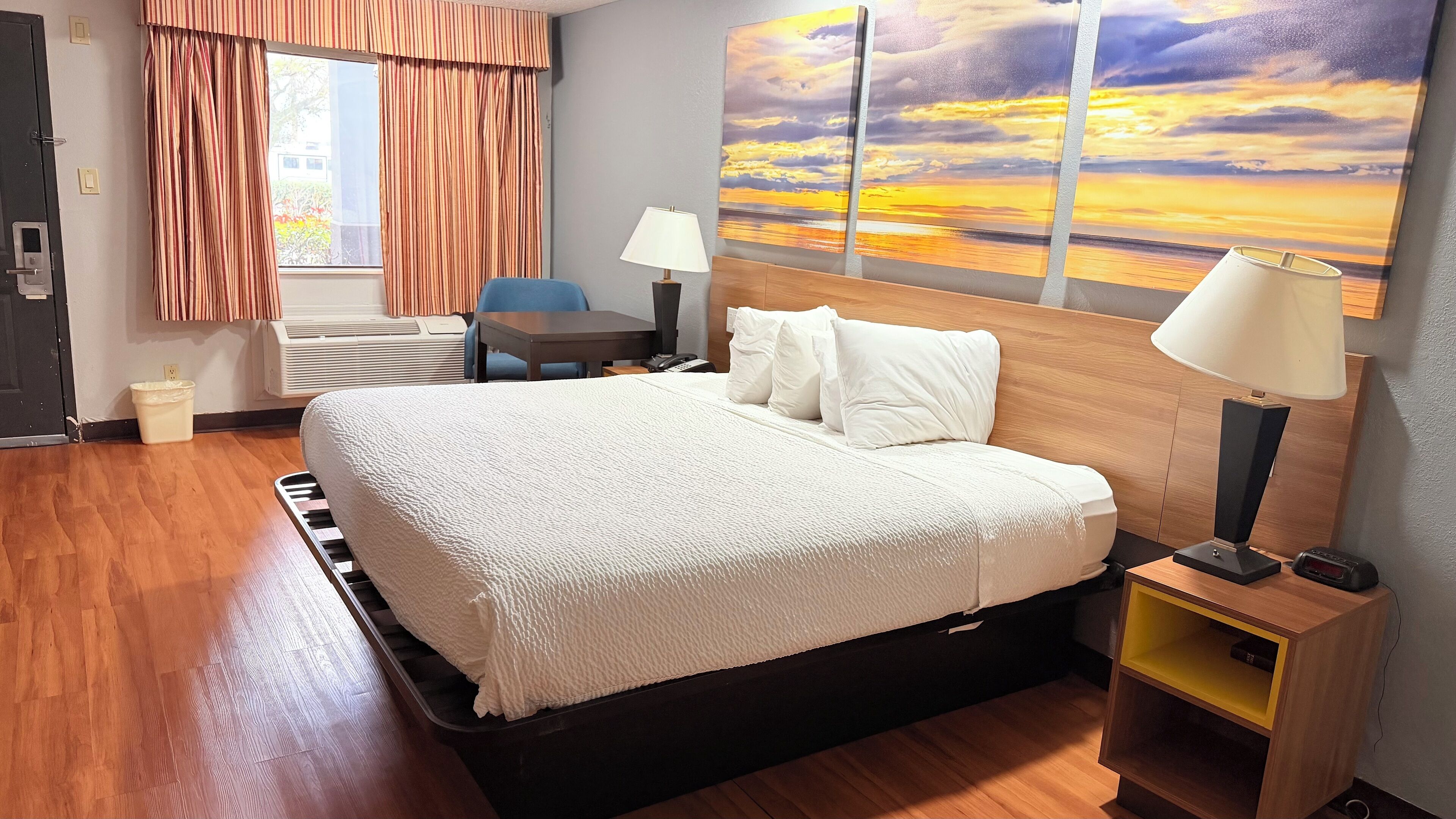 Habitación de hotel con una cama grande, un escritorio, una silla, una mesita de noche, una lámpara y un cuadro de un atardecer.