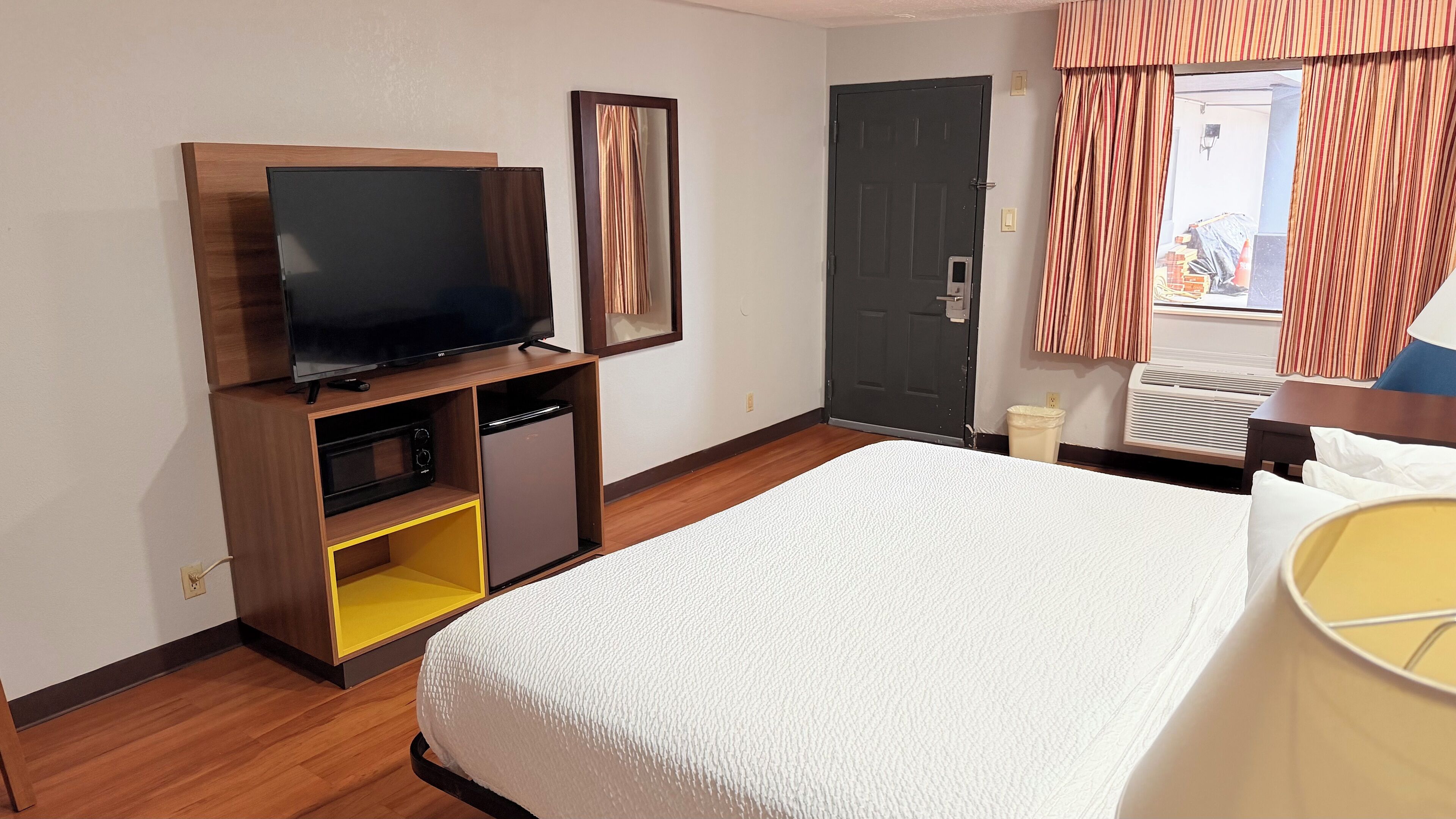 Une chambre d’hôtel avec un lit, un téléviseur posé sur un meuble en bois, un petit réfrigérateur, une porte et une fenêtre avec des rideaux.