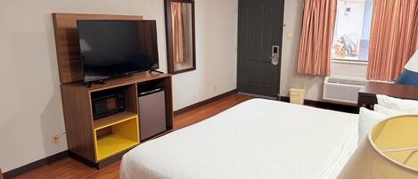 A hotel room with a bed, a television on a wooden stand, a small refrigerator, a door, and a window with curtains.