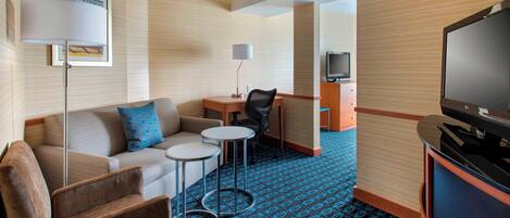 A hotel room with a sofa, armchair, small round tables, a desk with a chair, a television, and a patterned carpet.