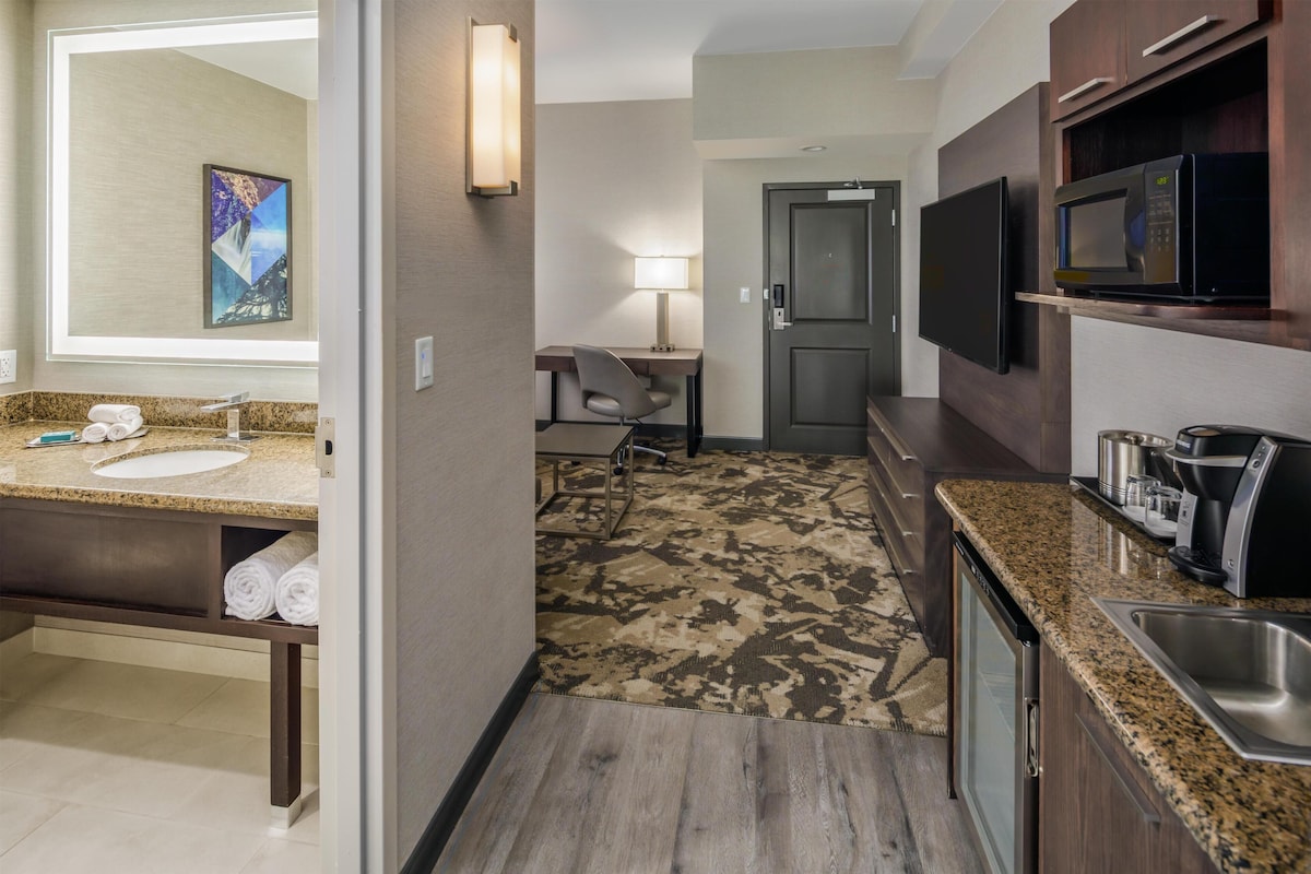 A hotel room with a bathroom, a desk, a chair, a television, and a kitchenette.