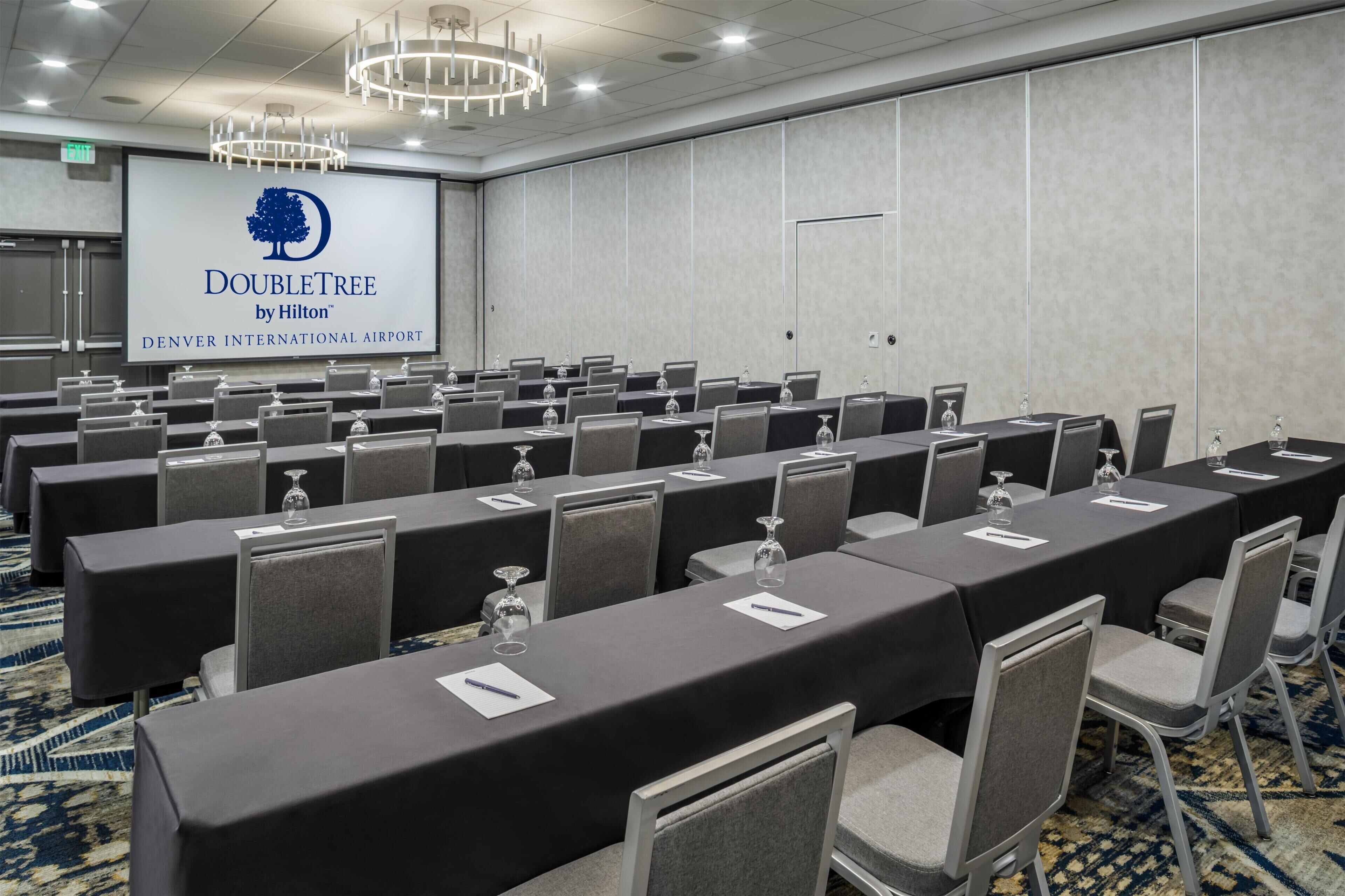 A conference room with rows of tables and chairs, a large screen displaying the DoubleTree by Hilton logo, and a carpeted floor.