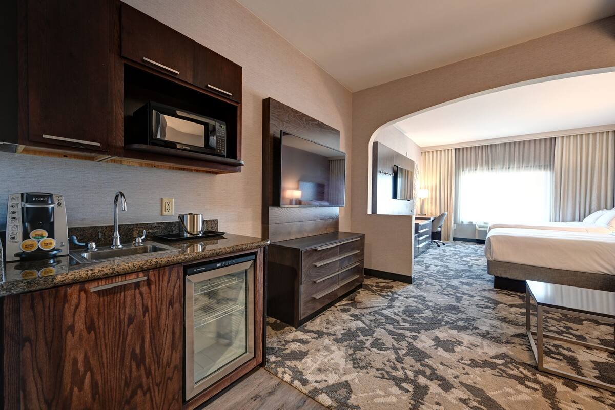 A hotel room with a kitchenette, a large bed, a TV, and a view of the city.