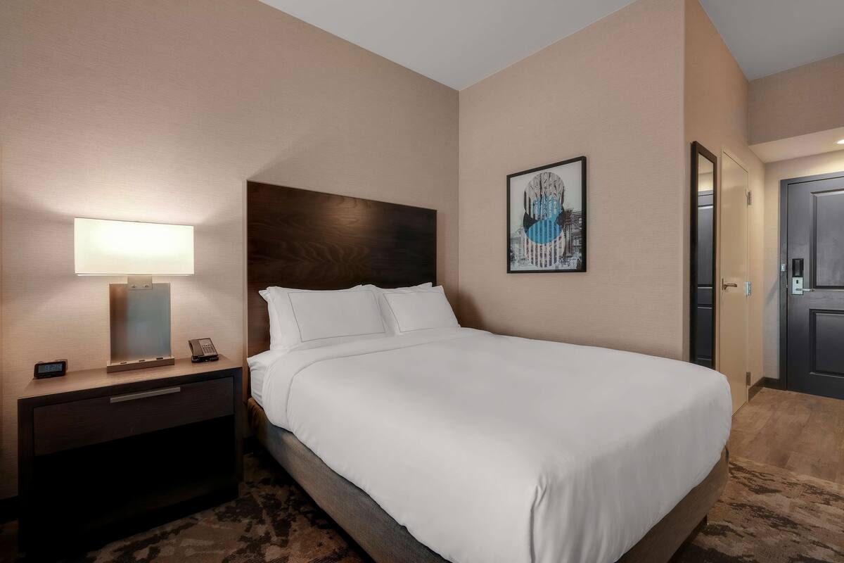 A hotel room with a bed, a bedside table with a lamp, a phone, and a painting on the wall.