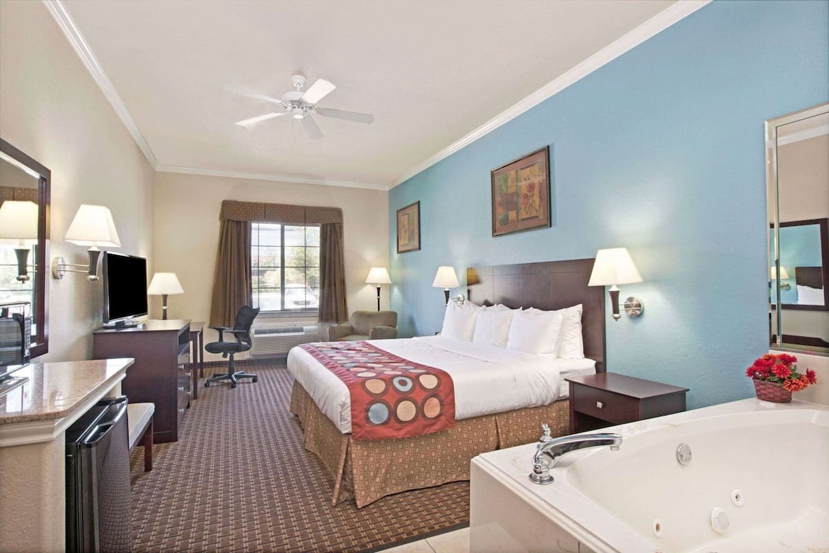 Une chambre d’hôtel comprenant un lit, un bureau, une chaise, une télévision, un ventilateur de plafond, une fenêtre et une salle de bain avec une baignoire.