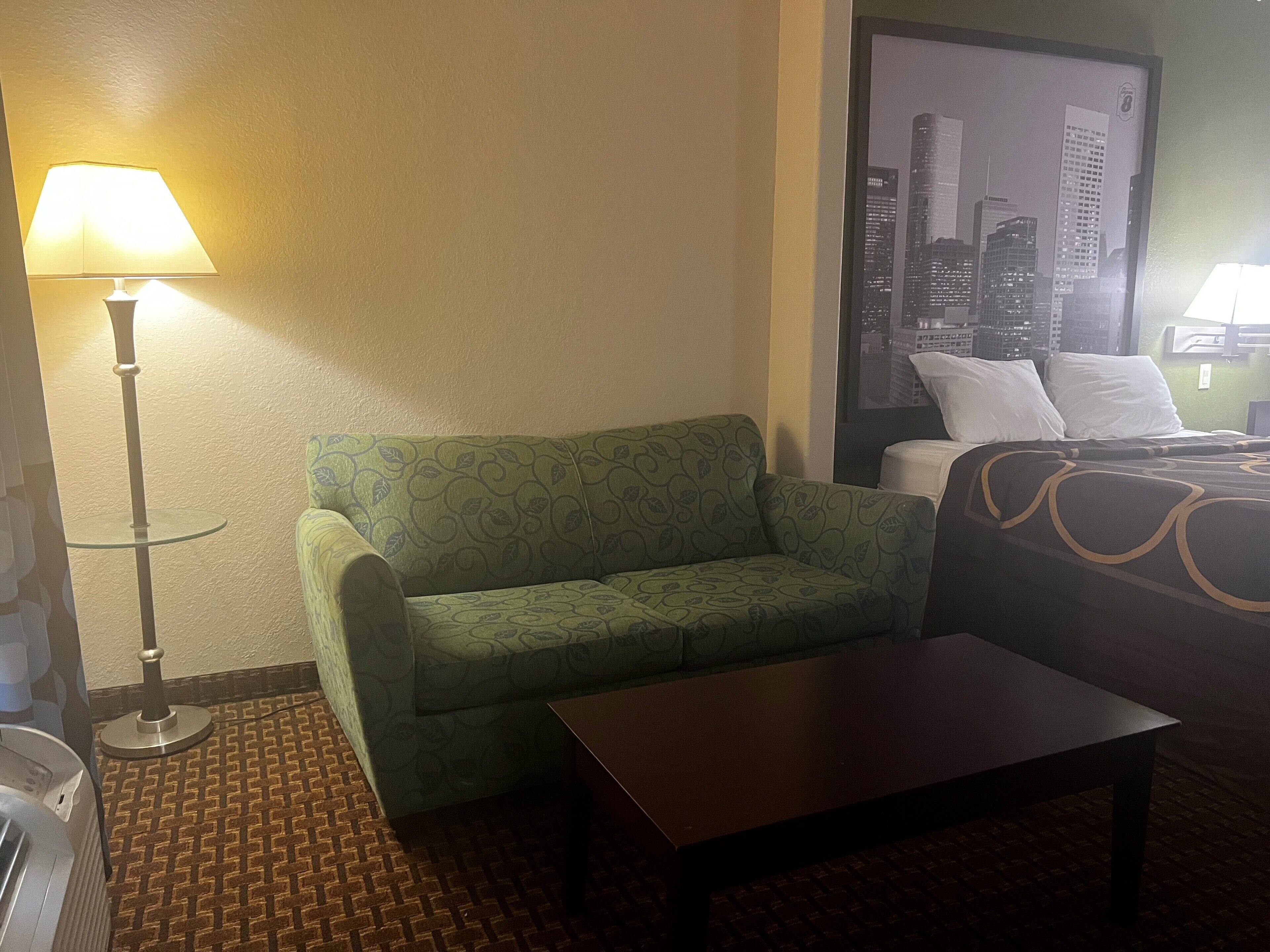 A hotel room with a green patterned sofa, a small table, a lamp, and a bed with a patterned bedspread.