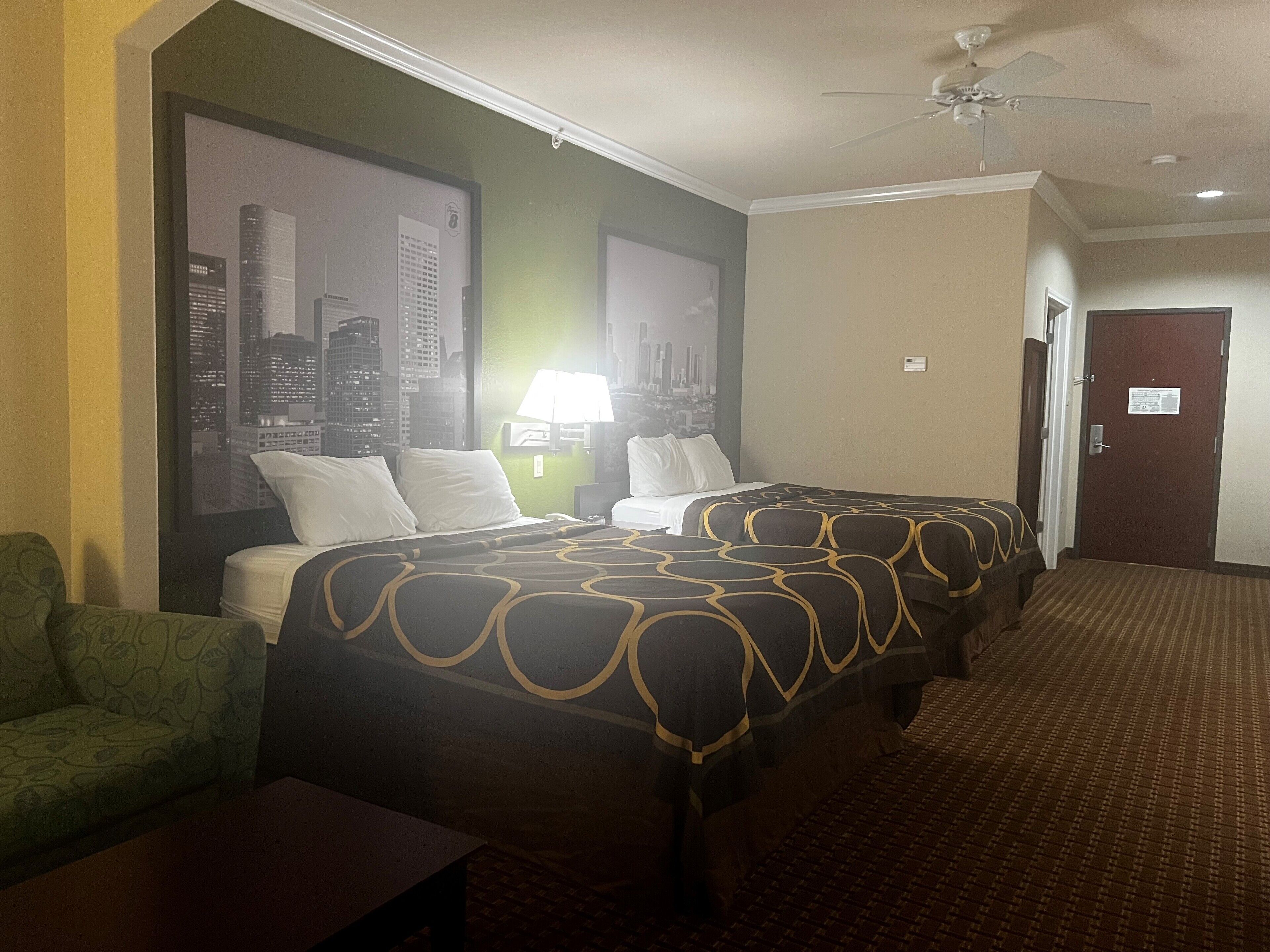 A hotel room with a bed, a green armchair, a nightstand, a lamp, and a cityscape view.