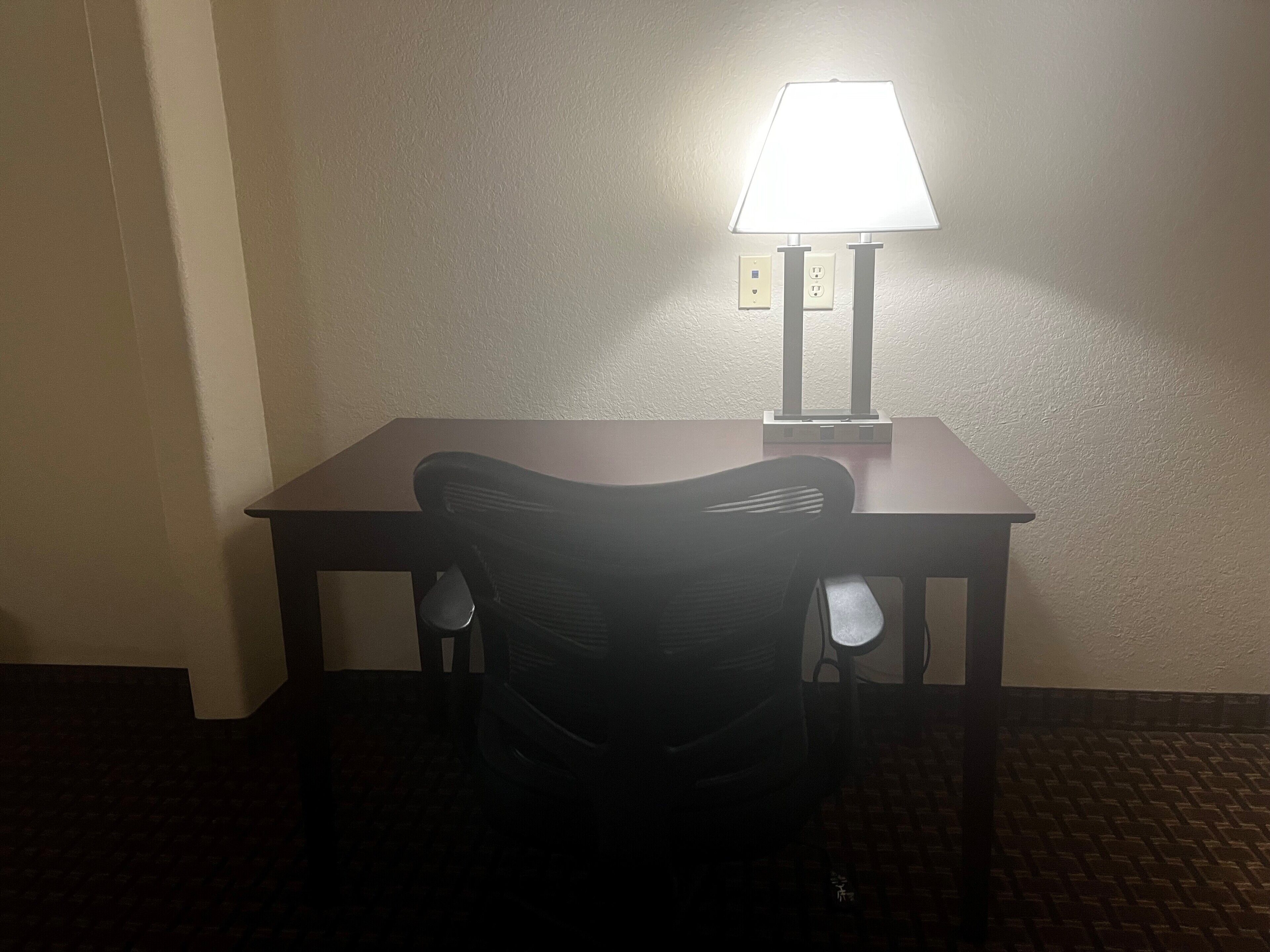 A hotel room with a single table and a chair, a lamp on the table, and a wall socket.