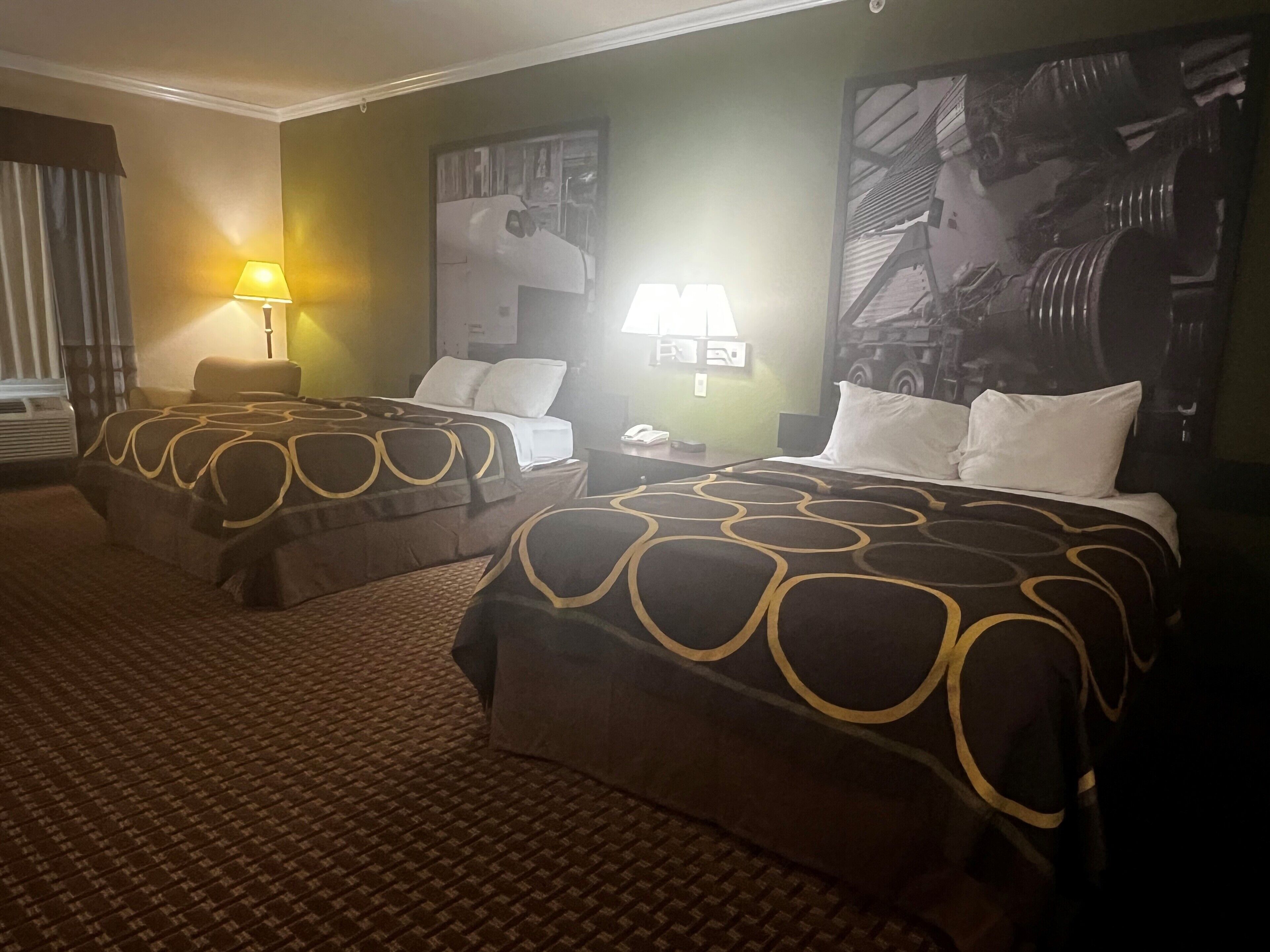 A hotel room with two beds, each with a patterned bedspread, a nightstand with a lamp, and a large abstract painting on the wall.