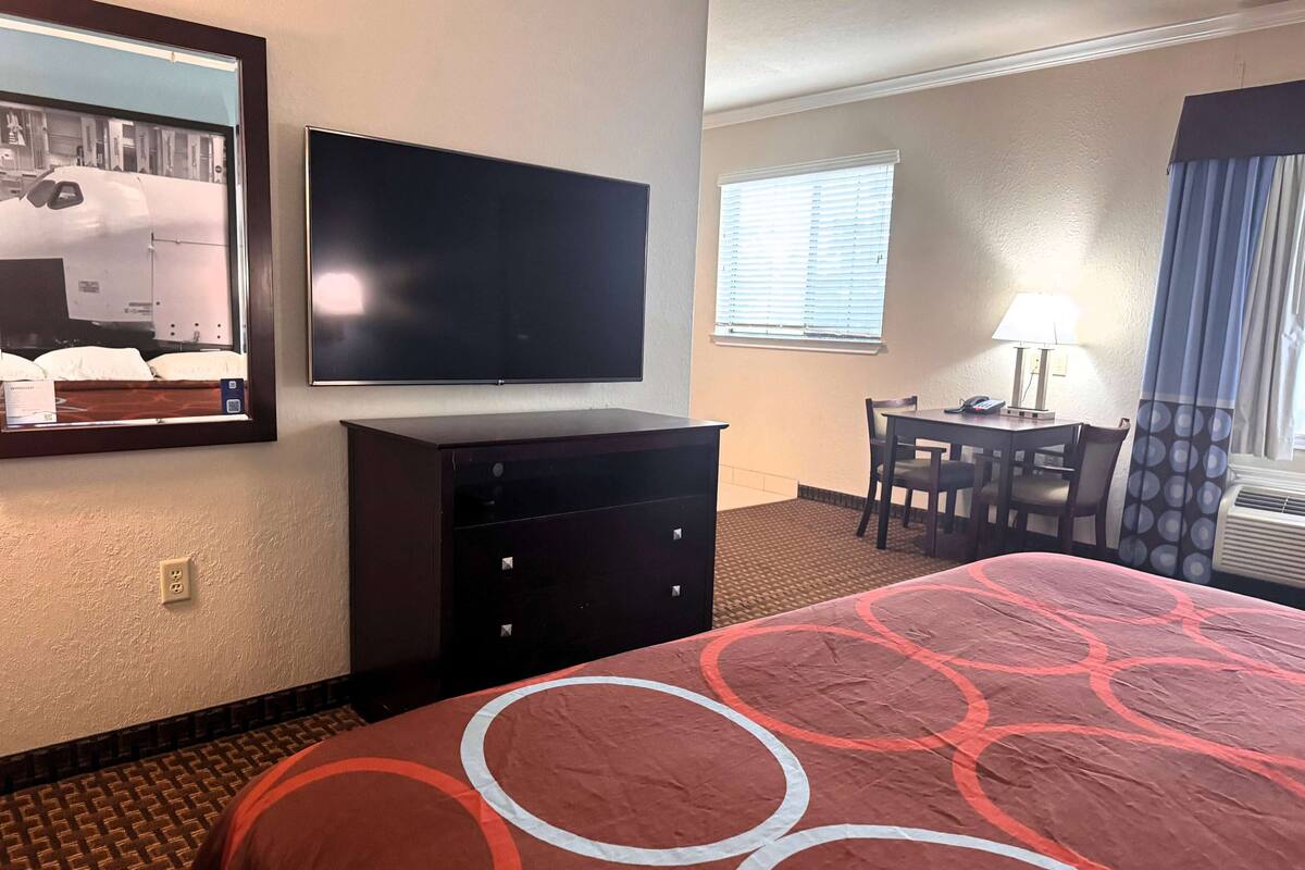 Une chambre d’hôtel comprenant un lit, un téléviseur fixé au mur, un bureau avec une chaise et une fenêtre avec des stores.