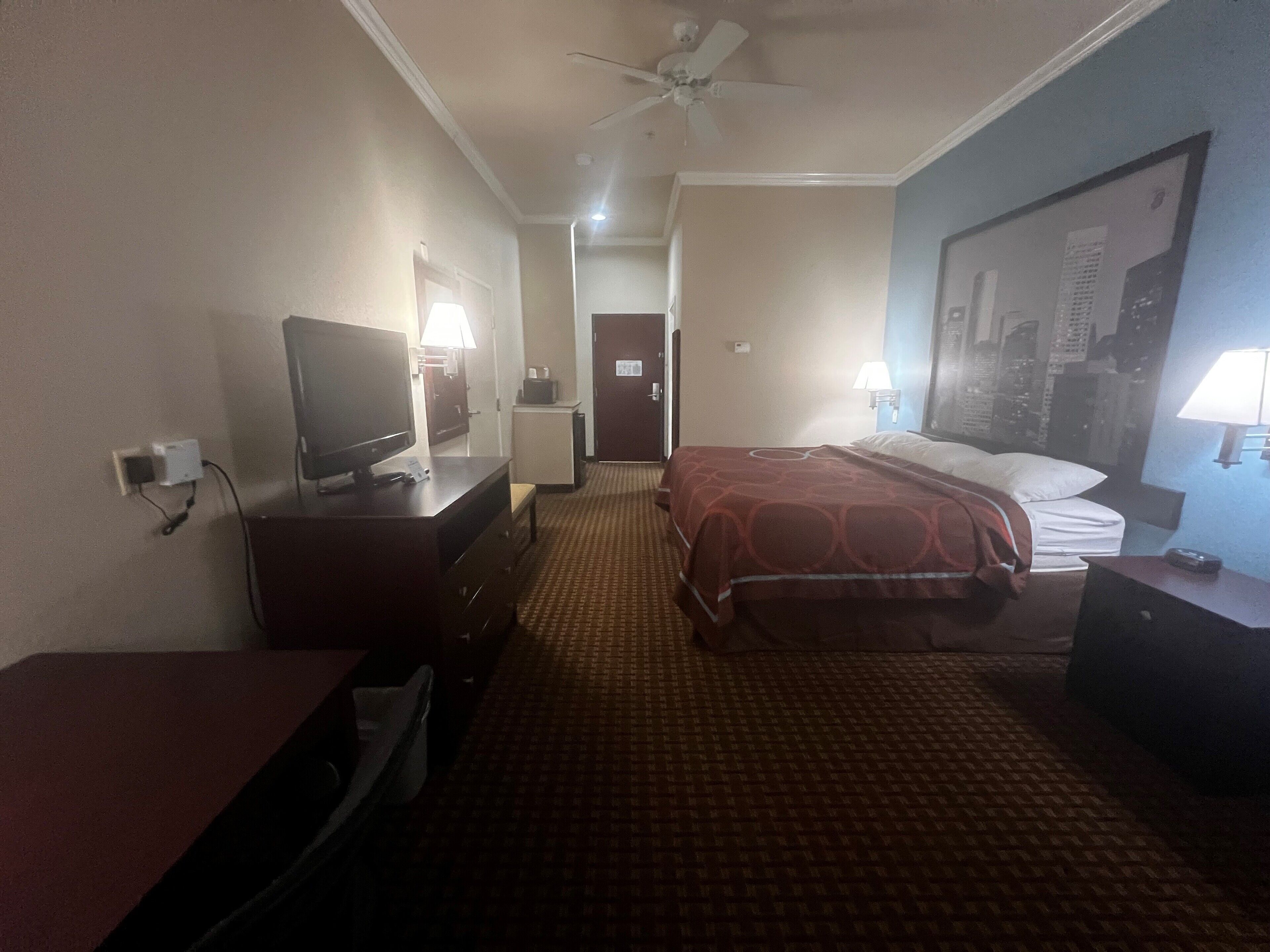 Une chambre d’hôtel comprenant un lit, un bureau avec une télévision, une lampe de bureau, une lampe fixée au mur et un ventilateur de plafond.