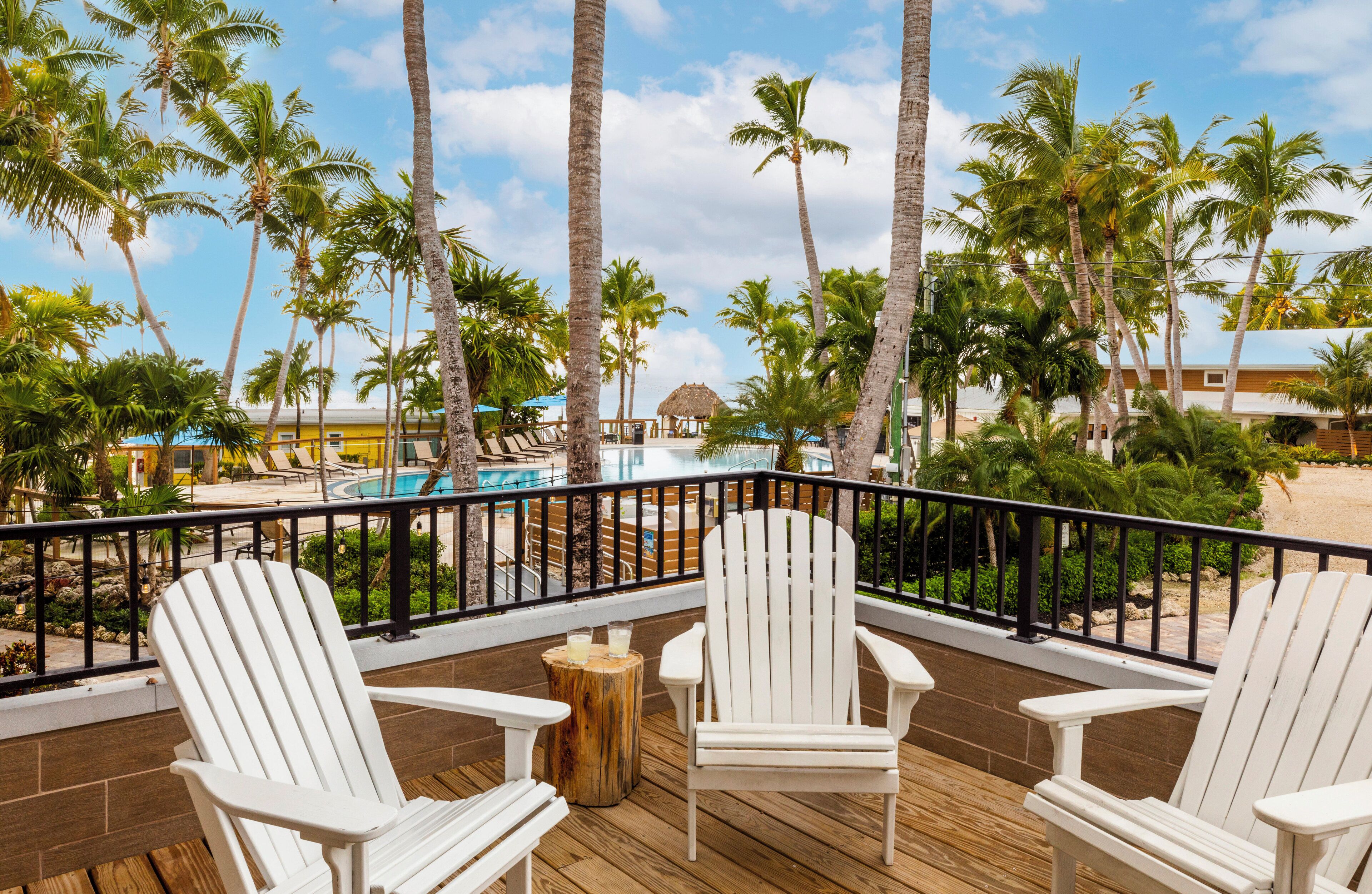 A wooden deck with white Adirondack chairs, a pool, and palm trees.