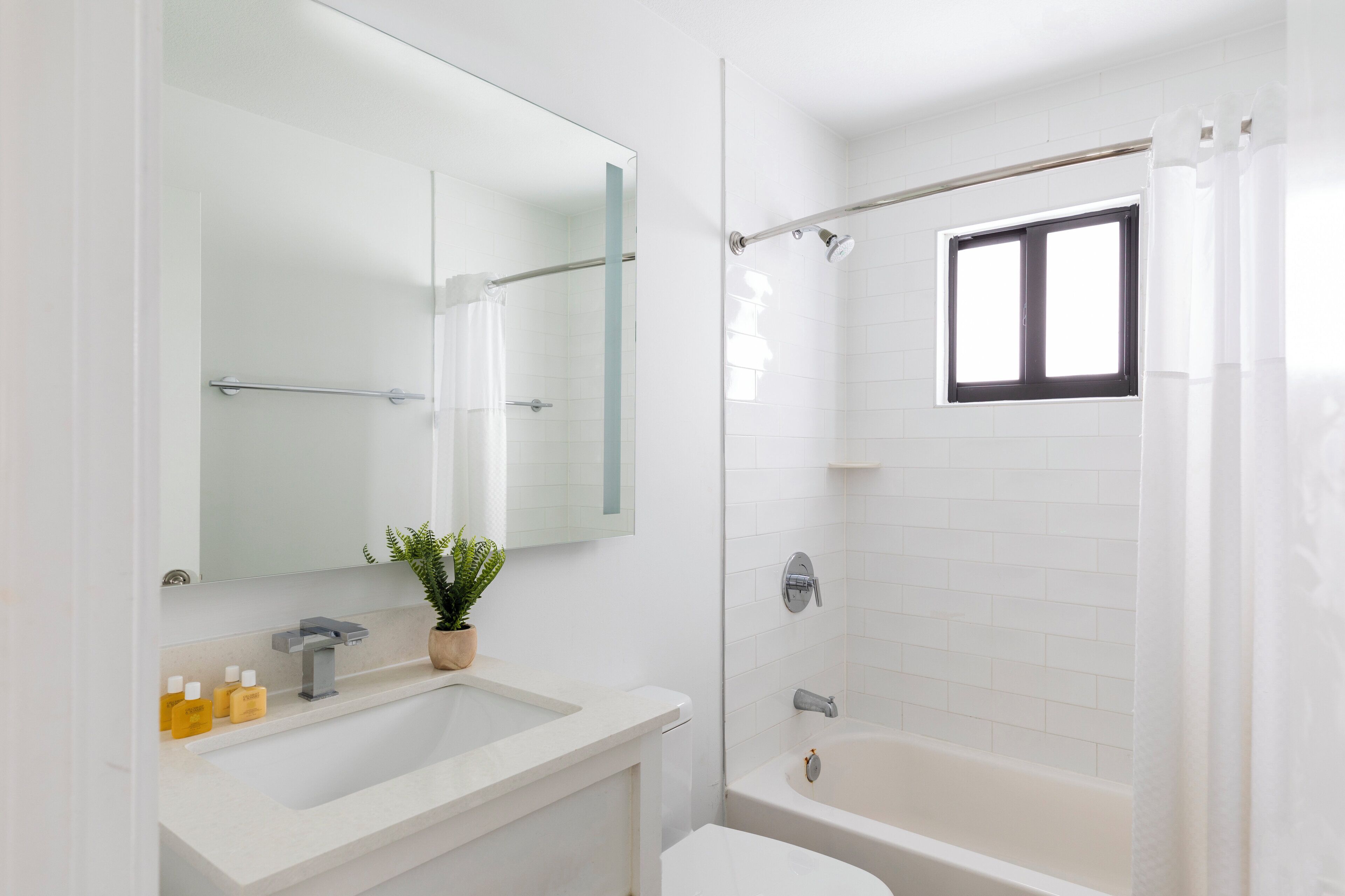 A modern bathroom with a white sink, a mirror, a shower, and a window.