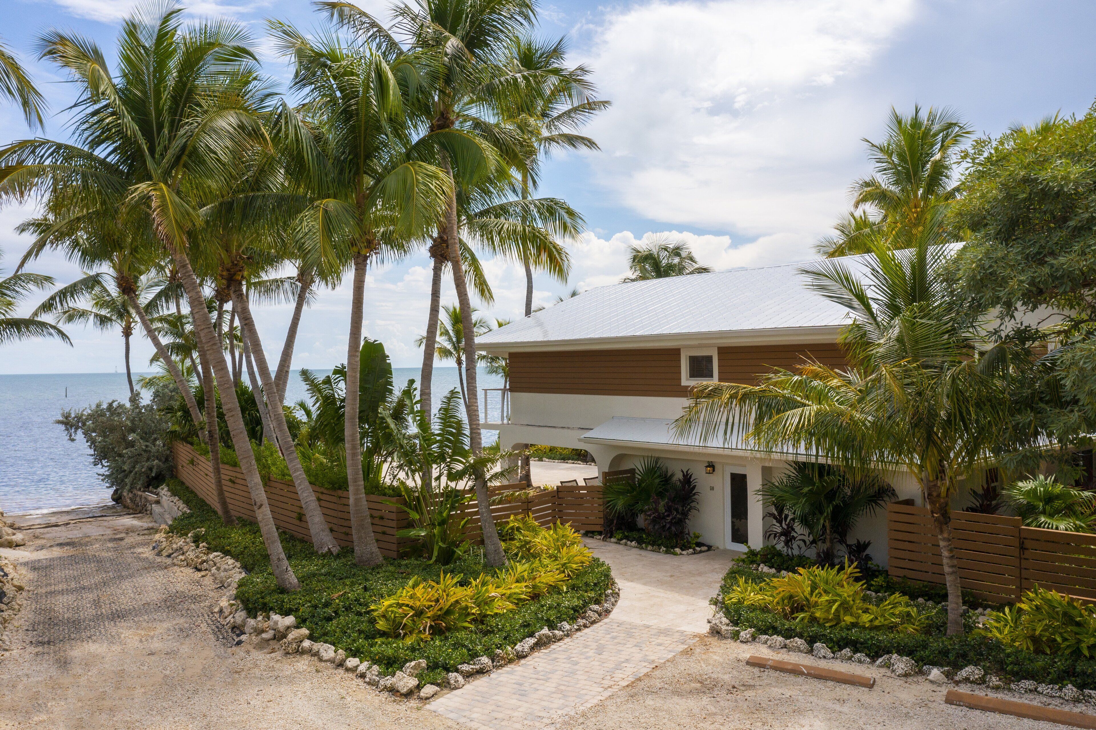 Una propiedad frente a la playa con una casa moderna, un camino de entrada pavimentado y una variedad de plantas tropicales.