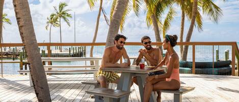 A wooden deck with a table and benches, surrounded by palm trees and overlooking a body of water.