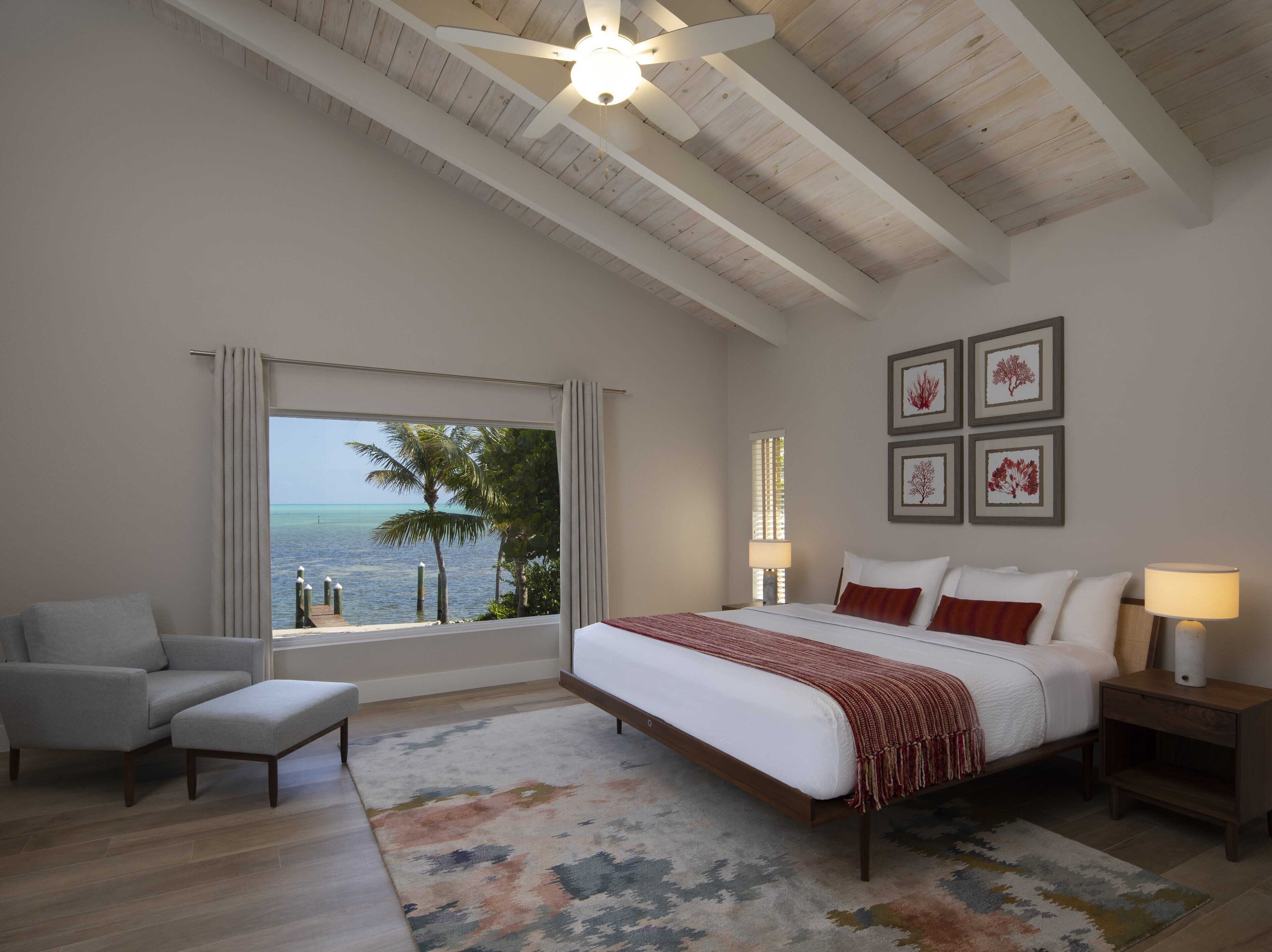 Une chambre avec un grand lit, une vue sur l’océan, un ventilateur de plafond et des œuvres d’art encadrées accrochées au mur.