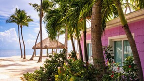 Um resort na praia com palmeiras, uma cabana de palha e um edifício roxo com uma janela.