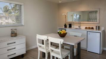 A modern kitchen with white cabinetry, a dining table with two chairs, a small refrigerator, and a window with blinds.