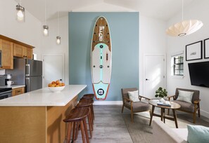 A modern kitchen with wooden cabinets, a white countertop, and a bowl of oranges. There is a surfboard mounted on the wall, a dining area with a table and chairs, and a living room with a sofa and a plant.