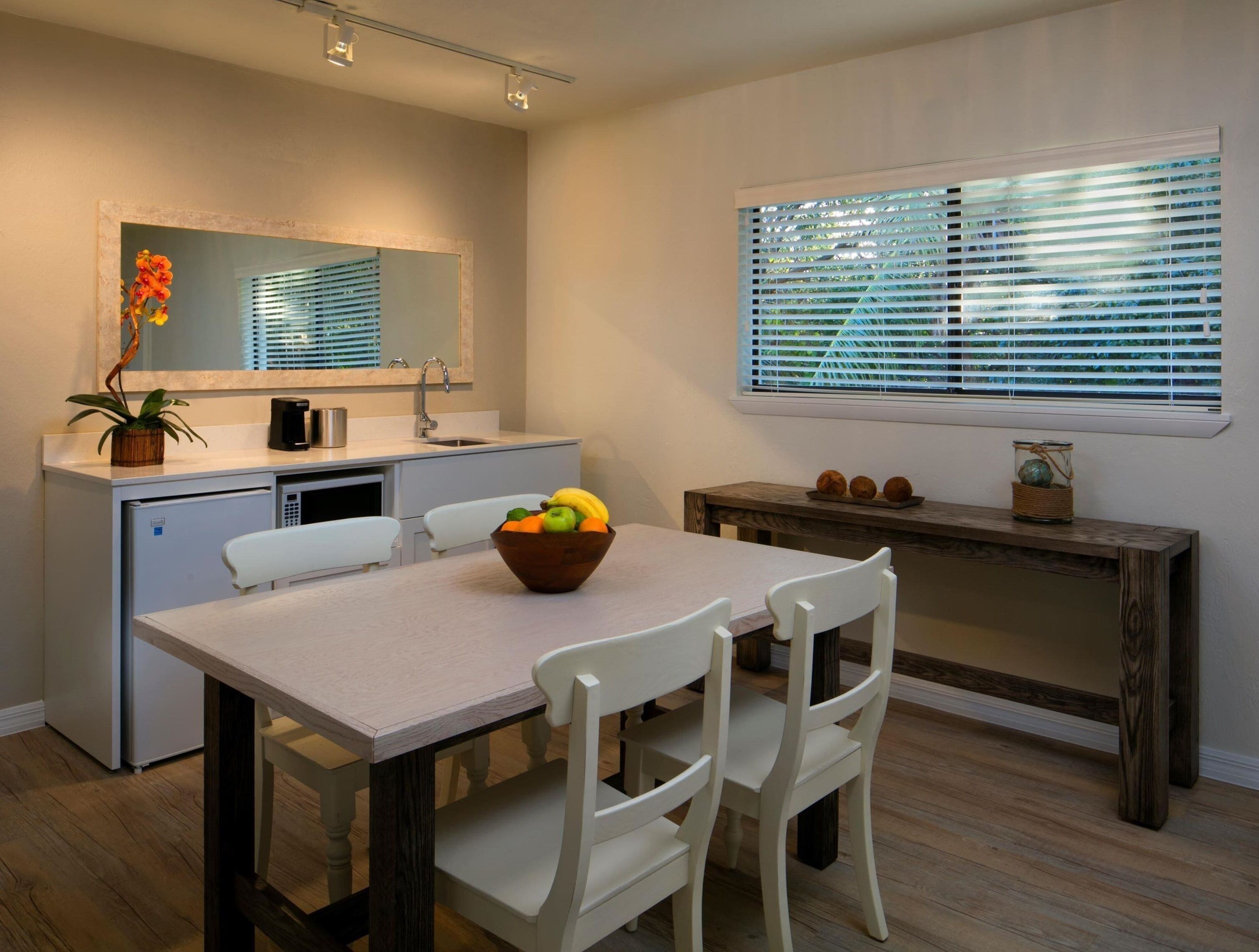 Une cuisine moderne avec un coin repas, comprenant une table en bois, des chaises blanches, un petit réfrigérateur et une fenêtre avec des stores.