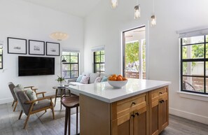 A modern kitchen with a large island, wooden cabinets, and a bowl of oranges. There are framed pictures on the wall, a television, and a dining area with chairs and a table.