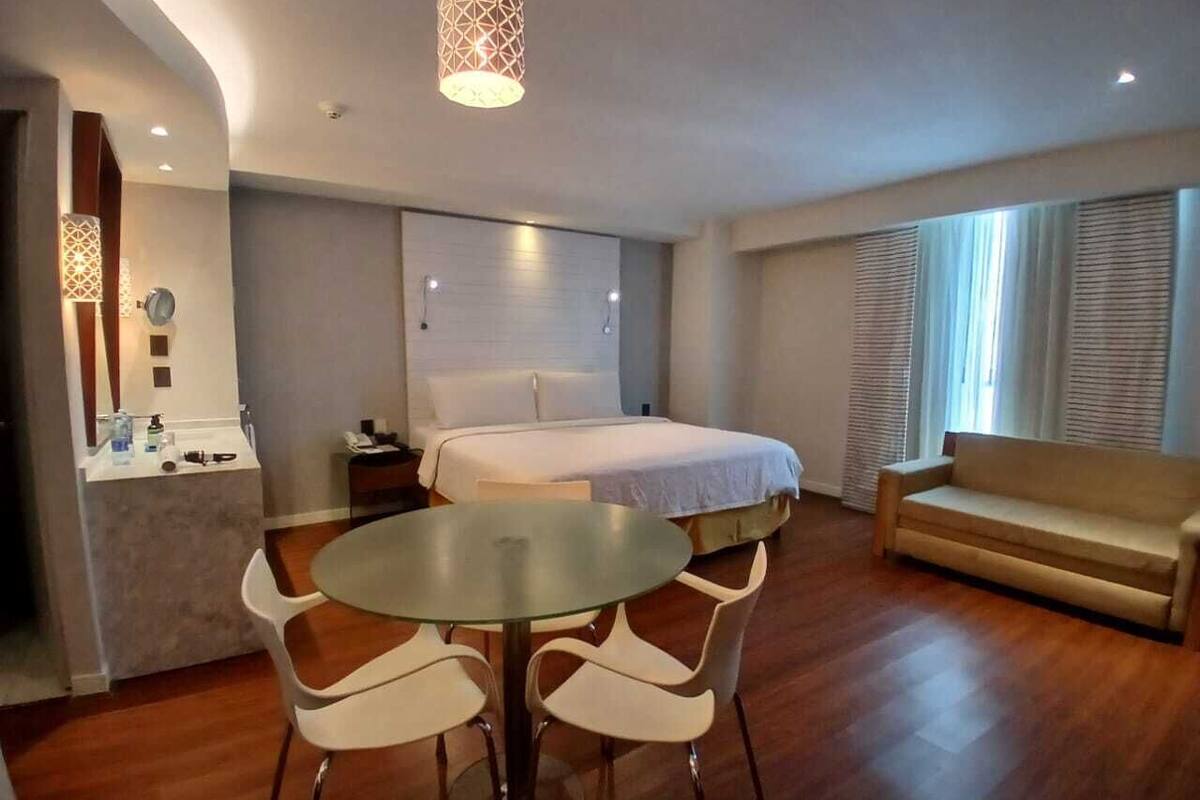 Ett hotellrum med en säng, en soffa, ett matbord med stolar, ett litet pentry och ett stort fönster med persienner.