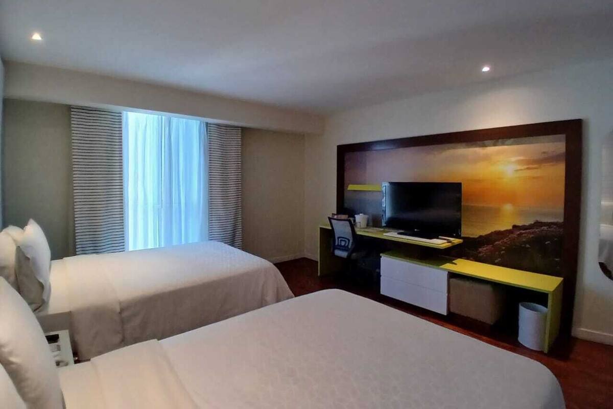 Ett hotellrum med två sängar, ett skrivbord med en tv och ett stort fönster med persienner.