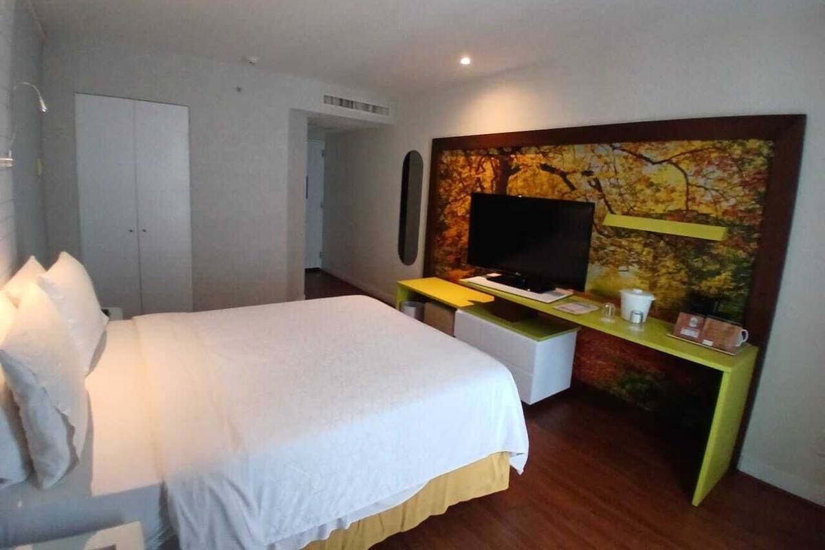 Ett hotellrum med en säng, ett skrivbord med en platt-TV, en kaffemaskin och en vägg prydd med en väggmålning med naturmotiv.
