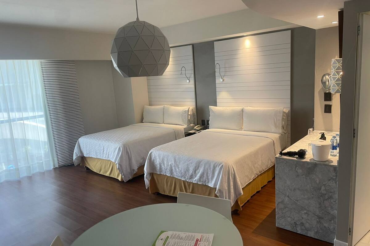 Ett hotellrum med två sängar, ett runt bord, ett skrivbord med en spegel och en hängande lampa.