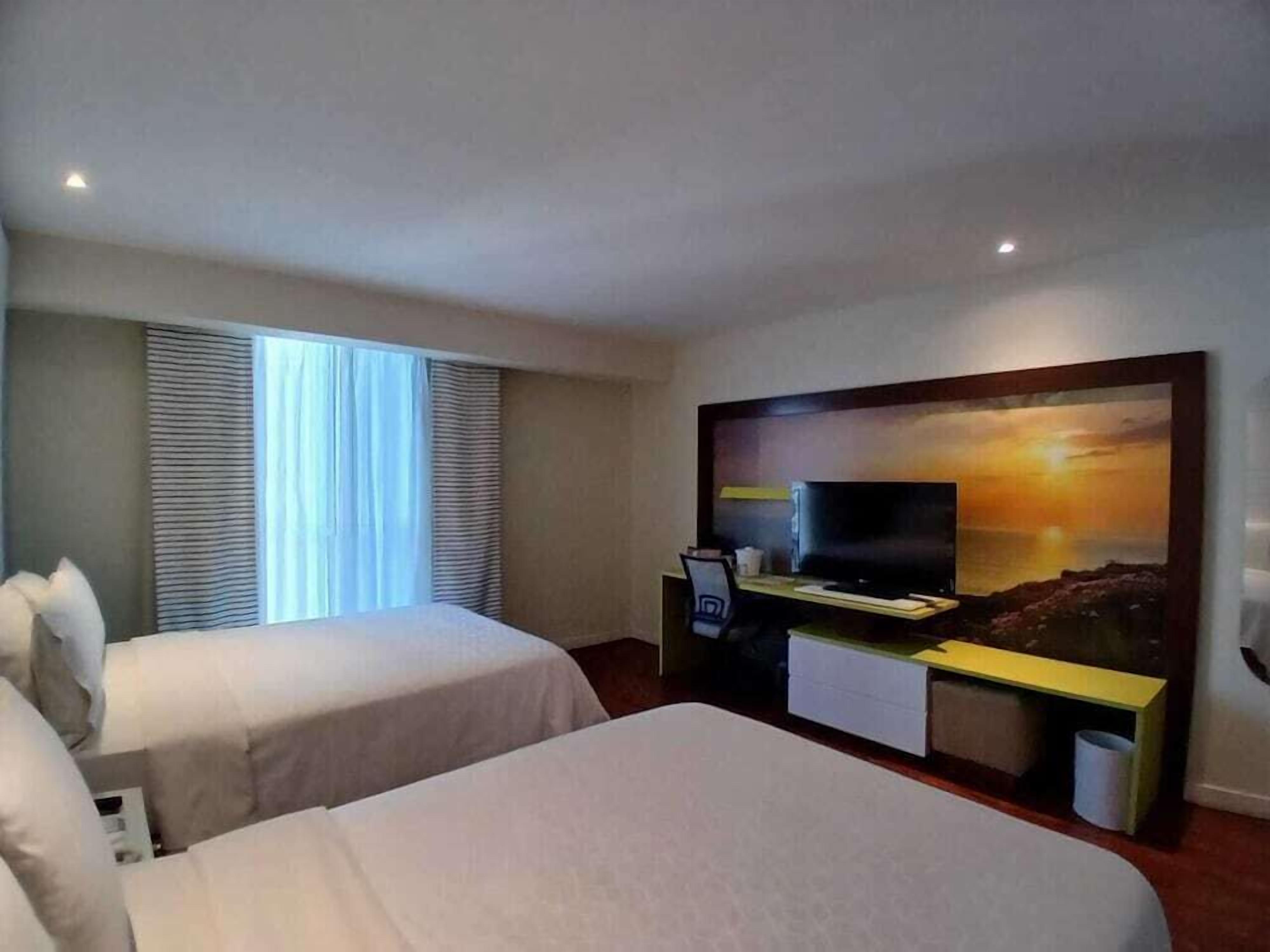 A hotel room with two beds, a desk with a computer, a TV, and a large painting of a sunset.