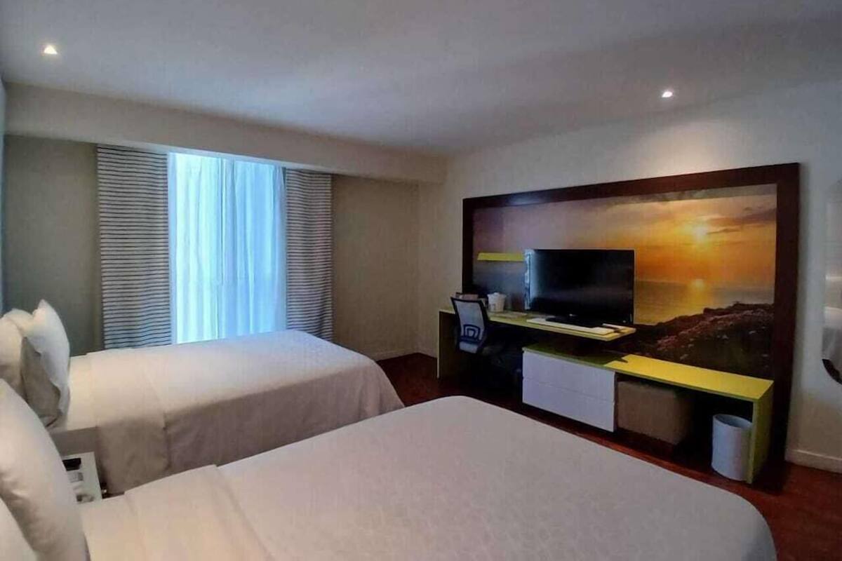 Ett hotellrum med två sängar, ett skrivbord med en dator, en tv och en stor tavla som föreställer en solnedgång.