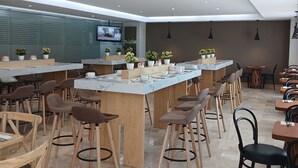 A modern cafe with multiple bar stools and tables, a television screen, and potted plants. - BelAir Business a Wyndham Garden Hotel (Mexico City)