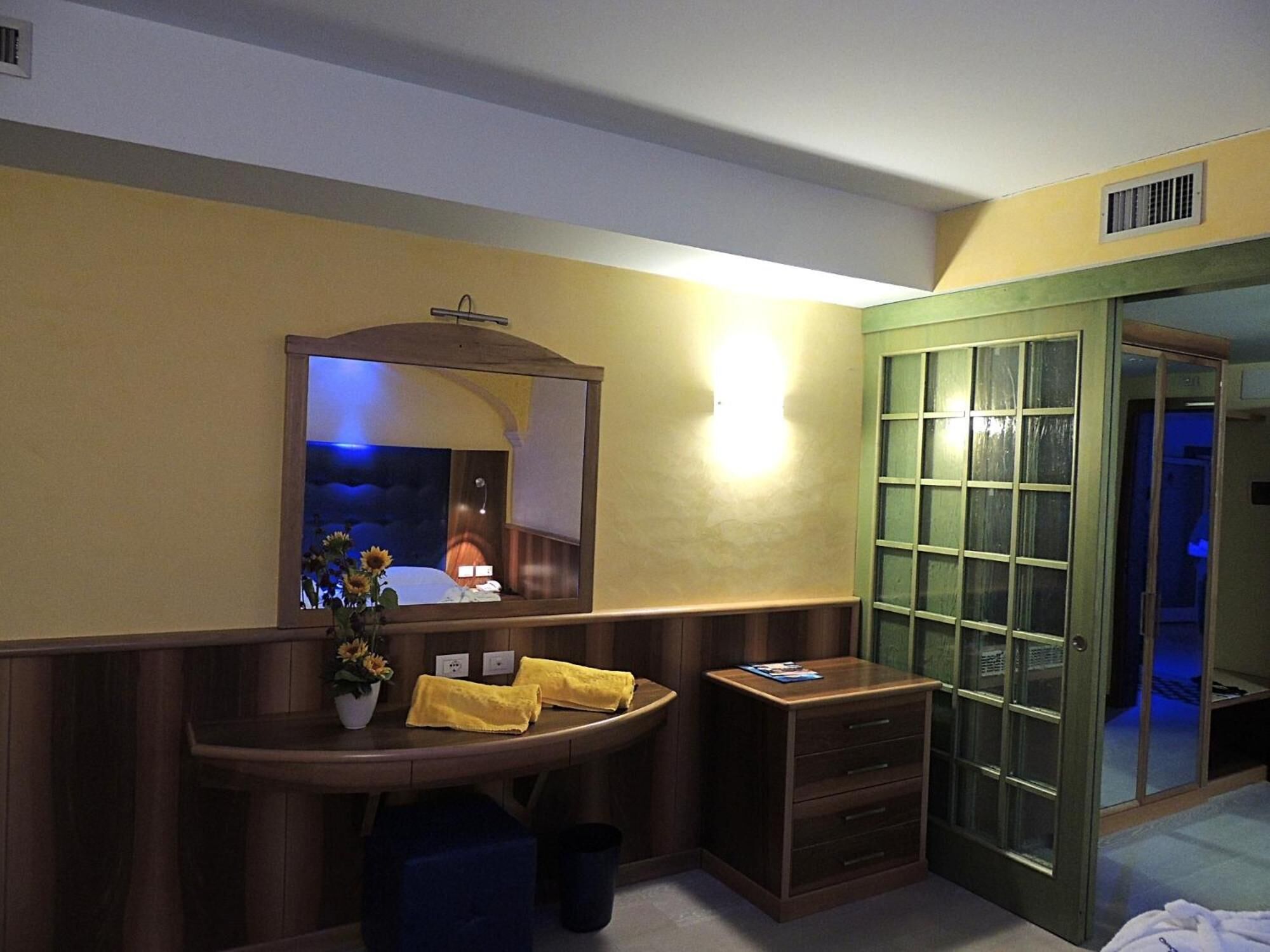 A hotel room with a wooden vanity, a mirror, a bedside table with a lamp, and a green door.
