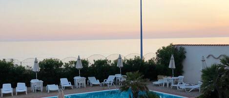 A pool area with lounge chairs and umbrellas, surrounded by greenery and a view of the sea.