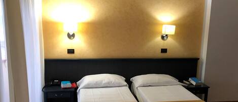 Une chambre d’hôtel avec deux lits simples, une table de chevet avec un téléphone et un objet rouge, et deux lampes fixées au mur.