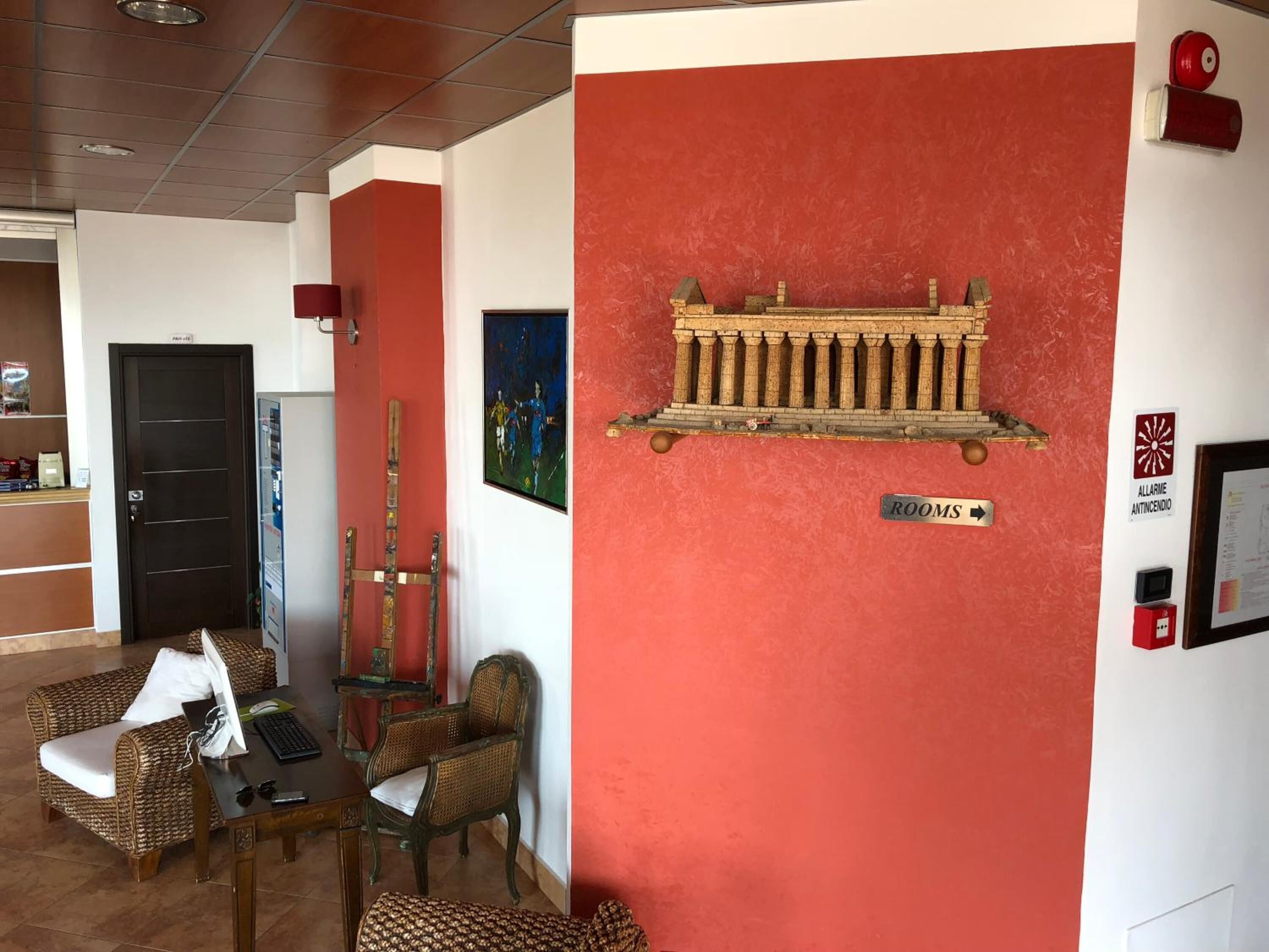 Een lobby met een rode accentmuur, een houten bank, een rieten sofa, een kleine tafel met een toetsenbord, en een bord waarop 'KAMERS' staat.