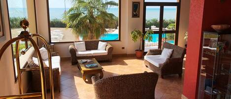 A spacious living area with wicker furniture, large windows offering a view of the beach and palm tree, and a glass cabinet with books.