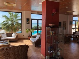 A spacious lounge area with wicker furniture, a glass display cabinet, and large windows offering a view of the pool and ocean.