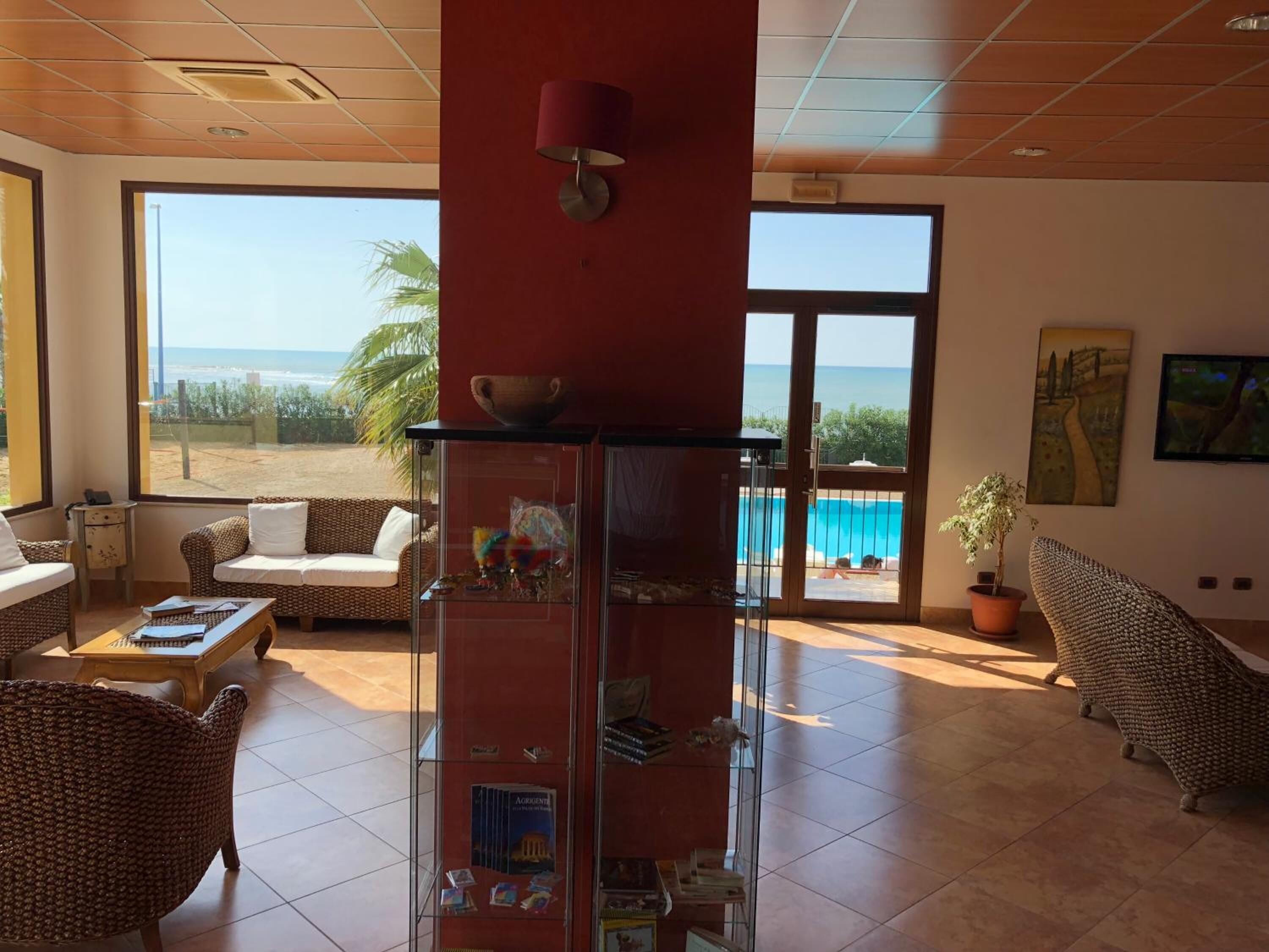 Amplia habitación con un gran ventanal que ofrece vistas a la playa y la piscina, mobiliario de mimbre y una vitrina de vidrio con libros.