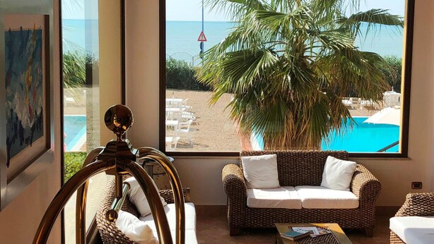 Habitación con un amplio ventanal con vistas a una piscina y a la playa, un sofá de mimbre y una silla decorativa.