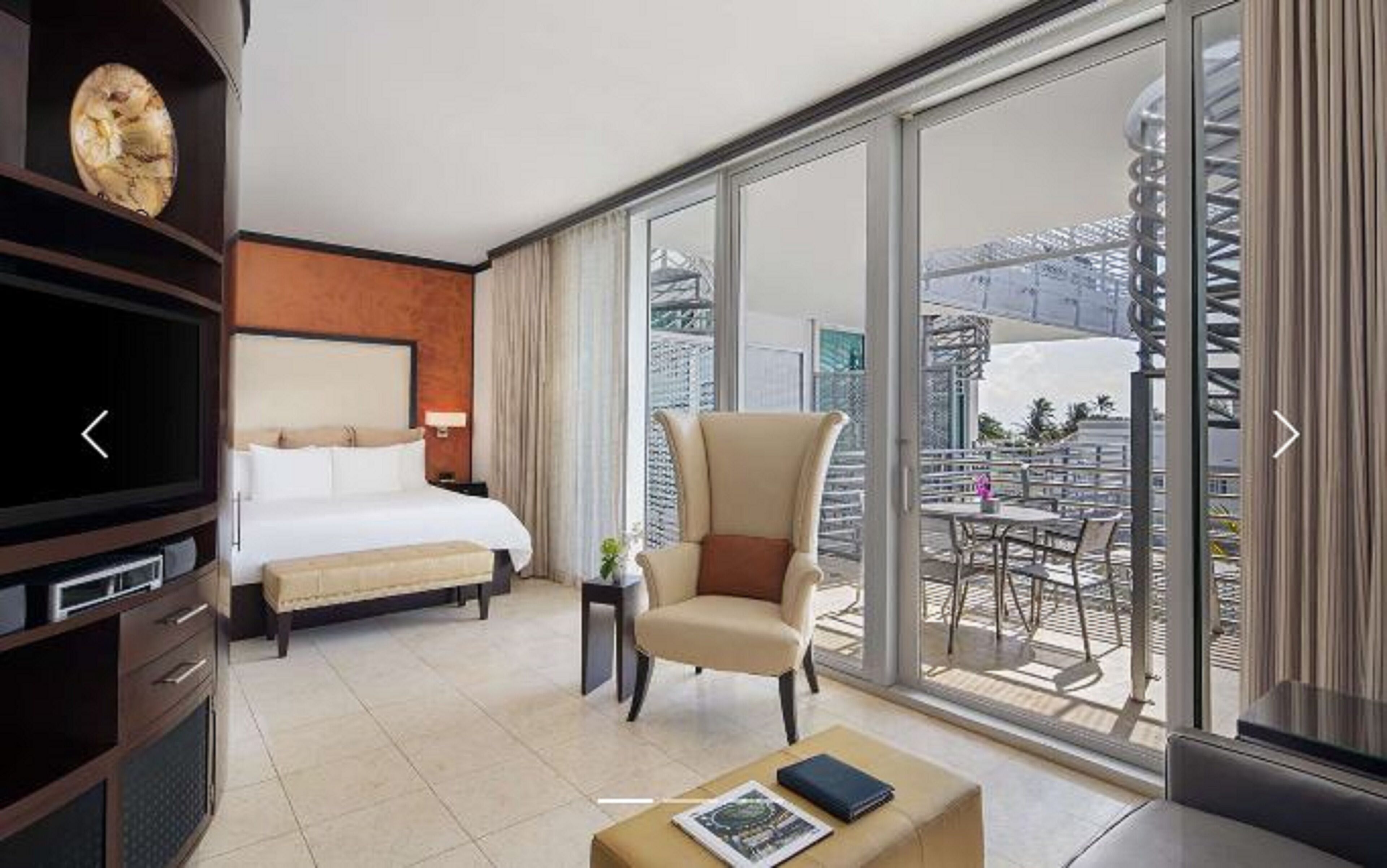 A hotel room with a bed, a chair, a TV, a coffee table, and a balcony with a table and chairs.