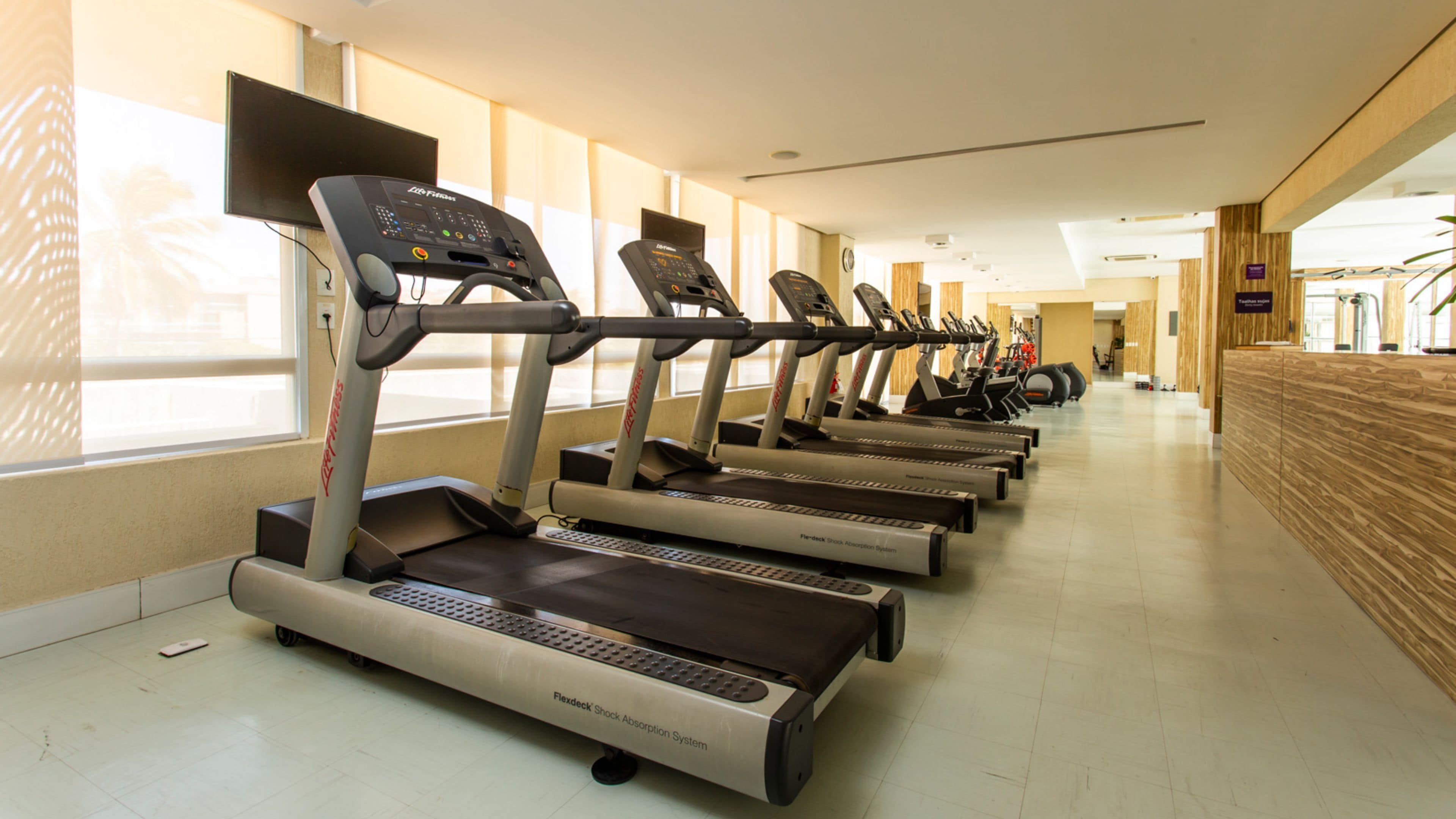 A row of treadmills in a well-lit indoor gym with large windows and a wooden reception desk.