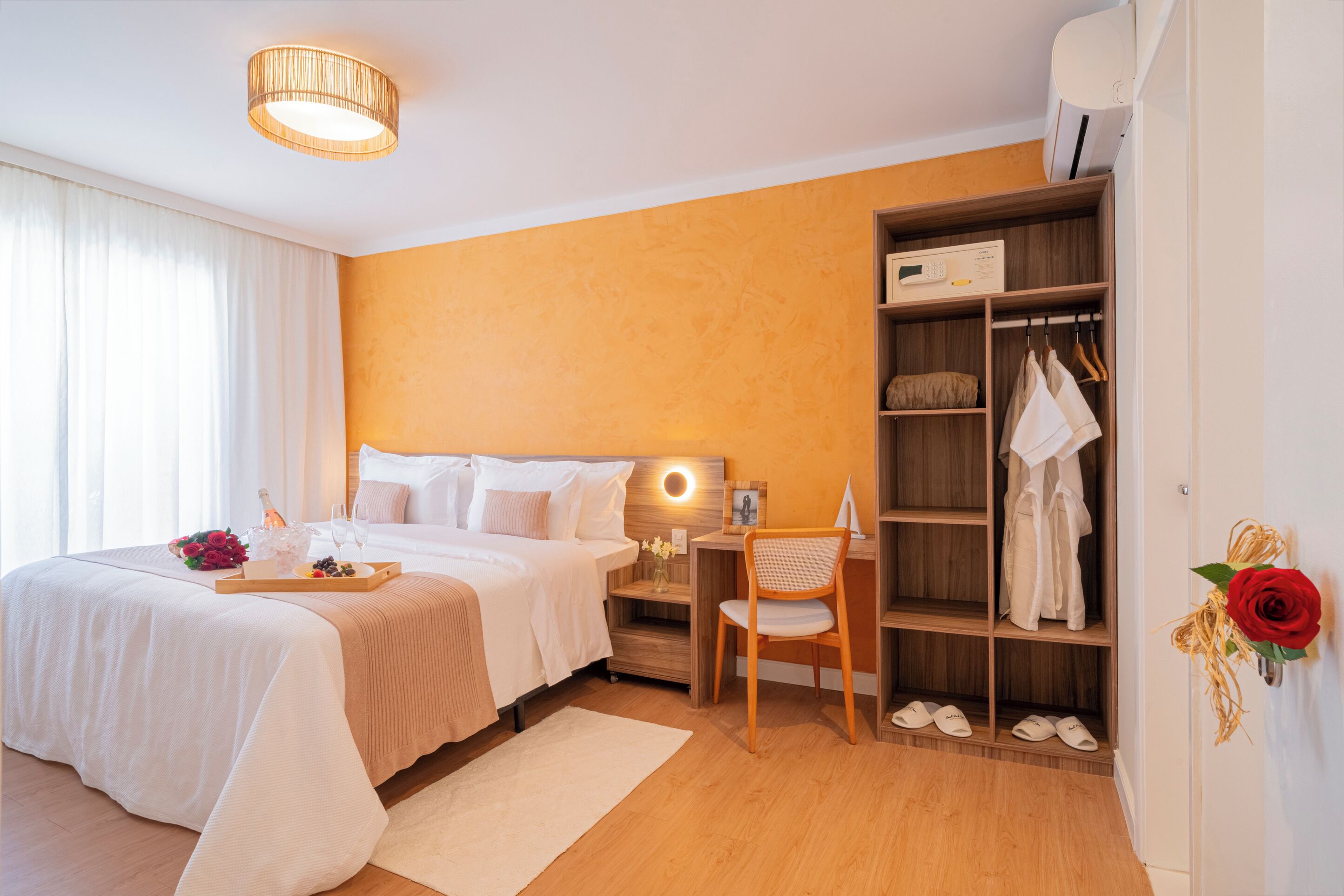 Quarto de hotel com cama, mesa de cabeceira, escrivaninha, cadeira, guarda-roupa e uma rosa vermelha decorativa.